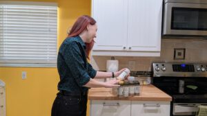 A professional home organizer in a kitchen. They are placing a bottle of spices into a container.